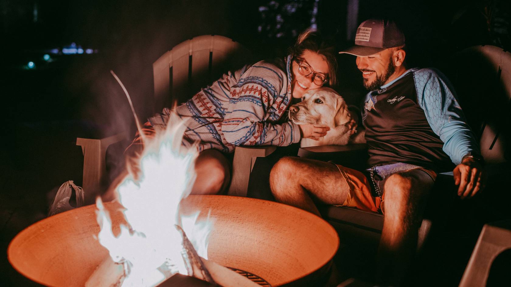 two people and a dog sitting on lawnchairs in front of a fire